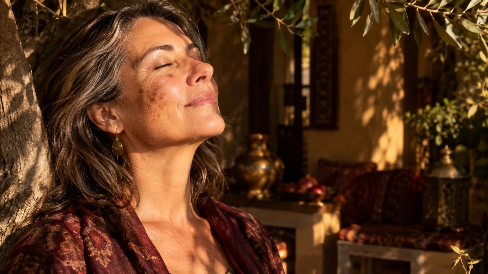 A woman in her fifties with sun spots on her cheek tilts her face toward warm dappled light, eyes closed.